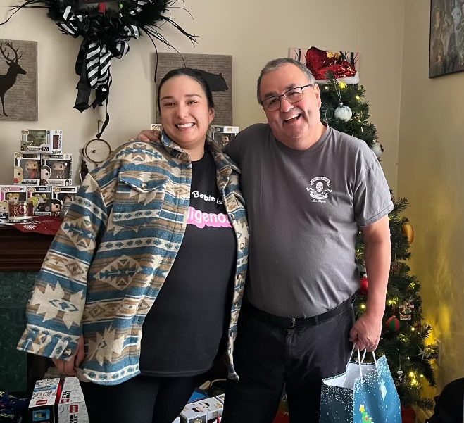 John G. Paul with Kendall (MSW’23), a mental health counsellor at the Mi’kmaw Native Friendship Centre. Paul and his adult child stand together in front of a Christmas tree holding gifts.