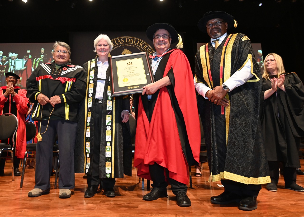 Paul accepting his honorary degree. (l-r) Elder Tom Christmas, President and Vice-Chancellor Dr. Kim Brooks, John G. Paul, Chancellor Dr. Rustum Southwell. Paul smiling holds his framed degree on the Convocation stage alongside members of the Dalhousie administration and faculty, all wearing ceremonial robes and garments.