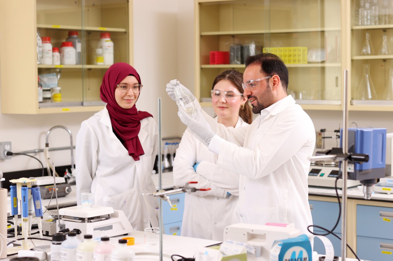 Two women and a man in white lab coats examine a glass beaker in a laboratory